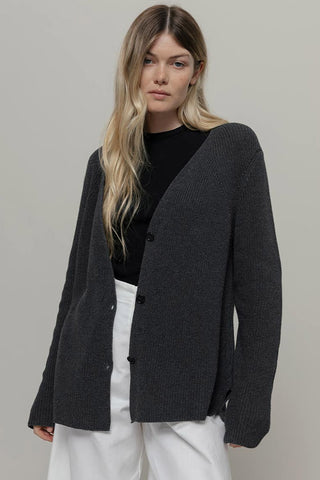 Woman wearing a loose fit dark grey cardigan with black top and white pants, in front of plain background. 