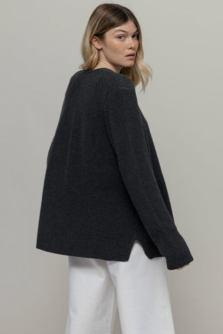 Woman looking back over her shoulder wearing loose fitting dark grey cardigan and white pants. 