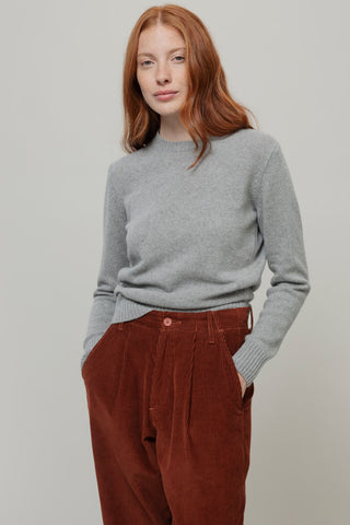 A woman with red hair wearing a light gray knit crewneck sweater and rust-brown corduroy trousers, standing against a neutral light gray background.