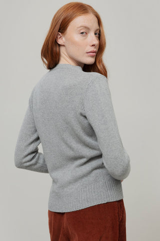A woman with red hair is shown from the back wearing a light gray knit sweater and rust-brown corduroy pants against a plain light gray studio background.