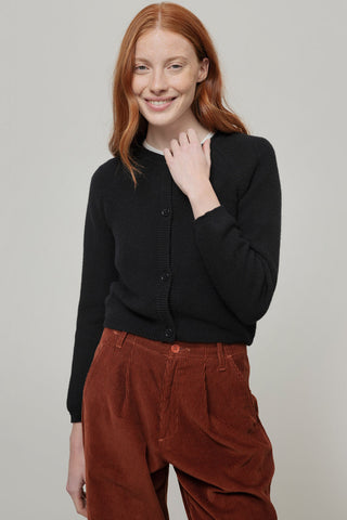 Model with red hair wearing a black knit cardigan with rust coloured corduroy pants, standing in front of grey background. 