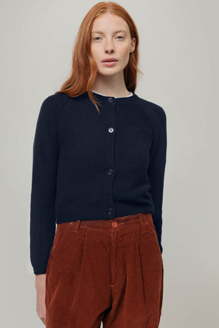A woman with red hair wearing a dark navy blue cardigan and rust-brown corduroy trousers, standing against a neutral light gray background.