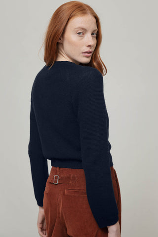 A woman with red hair is shown from a back-three-quarter angle wearing a dark navy ribbed knit sweater and rust-brown corduroy trousers against a plain light gray studio background.
