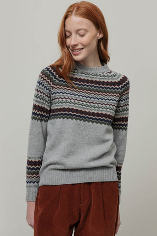 A woman with red hair wearing a gray knit sweater with a multicolor Fair Isle pattern across the chest and sleeves, paired with rust-brown corduroy pants, against a solid light gray background.