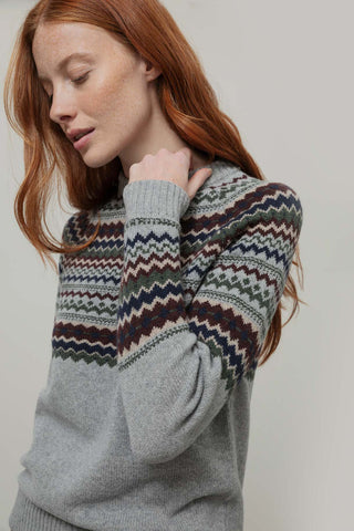 A woman with red hair wearing a gray knit sweater with a multicolor Fair Isle pattern across the chest and sleeves, posed against a neutral light gray background.