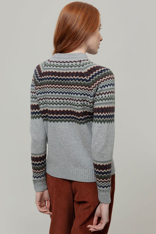 A woman with red hair is shown from the back wearing a light gray knit sweater with a multicolor Fair Isle pattern across the shoulders, upper back, and sleeves, paired with brown pants, against a solid light gray background.
