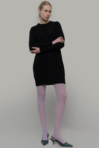 Woman in front of grey background wearing black recycled Selvaggia sweater dress by Rifo, paired with purple tights and green kitten heels. 