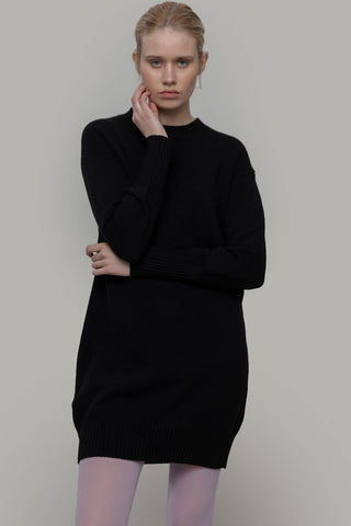 Woman in front of grey background wearing black recycled Selvaggia sweater dress by Rifo, paired with purple tights.