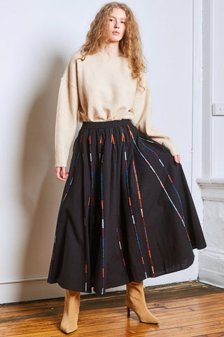 Woman in corner of a room with beige sweater, black ankle length skirt with colourful piping and tan suede heeled boots. 