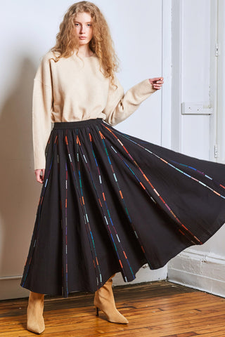 Model in corner of a room with beige sweater, black ankle length skirt with colourful piping and tan suede heeled boots. 