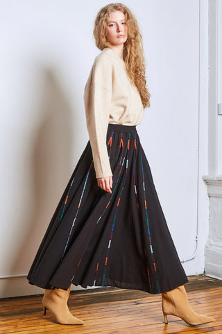 Model in corner of a room with beige sweater, black ankle length skirt with colourful piping and tan suede heeled boots. 