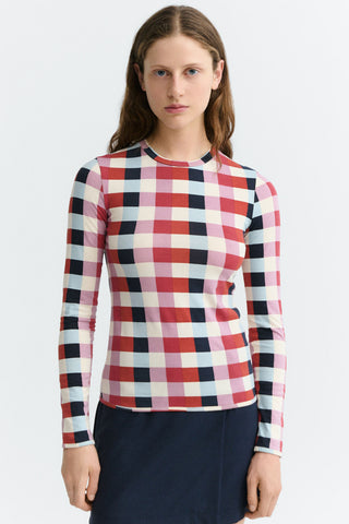 A woman wearing a long-sleeve fitted top top with a pink, red, black and white checkered pattern, paired with a dark skirt, against a light gray studio background.