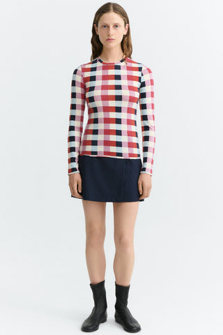 A model stands against a light gray studio background wearing a long-sleeve top with a red, pink, black, and white check pattern, paired with a dark black mini skirt and black ankle boots.