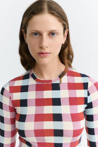 A woman wearing a long-sleeve crewneck top with red, pink, black and cream check print, set against a plain light gray background.