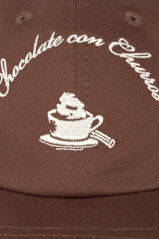 Close-up of a brown baseball cap with white embroidered text reading 'Chocolate con Churros' and a white coffee cup on the front.