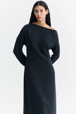 A woman wearing a long-sleeve, off-the-shoulder black dress against a plain white studio background.
