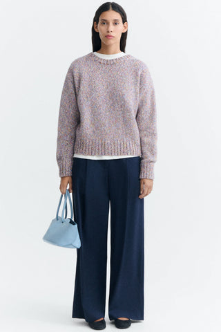 A model stands against a plain white studio background wearing a multi-coloured speckled sweater, dark navy wide-leg trousers, black flats, and holding a light blue handbag.