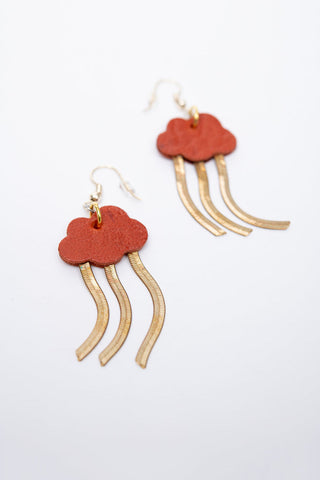A pair of cloud-shaped drop earrings with coral leather cloud with three dangling gold-tone chains, shown on a clean white background.