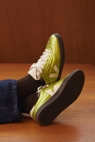Person shown from calves down wearing metallic lime green Veja sneakers with brown socks against wood grain background. 