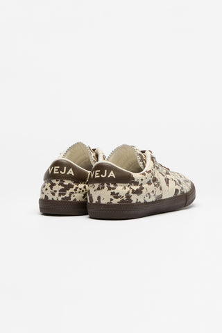 A pair of low-top sneakers shown from the back, featuring a beige and brown camouflage pattern with dark brown soles and 'VEJA' branding on the heel.